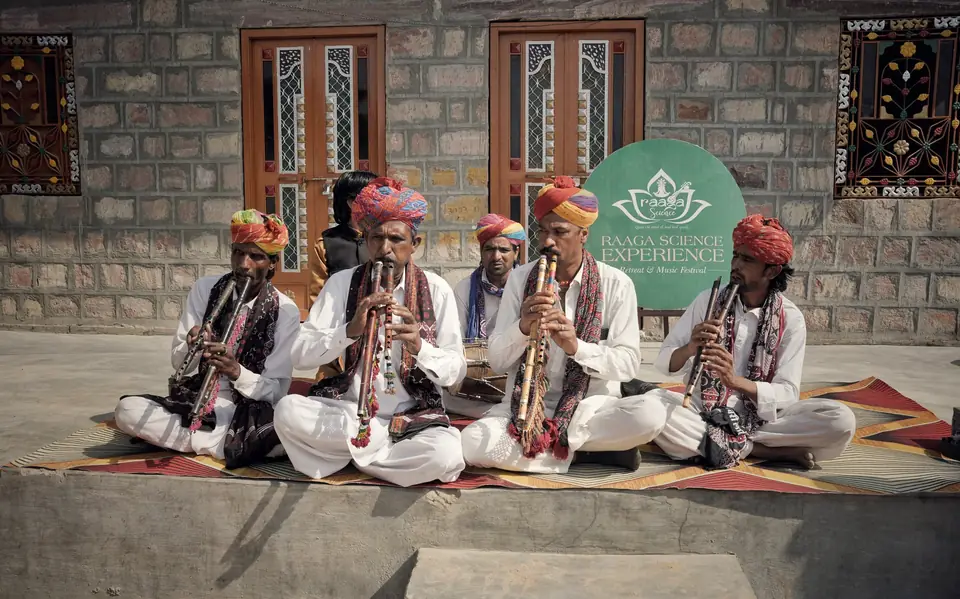 Folk musicians in turbans seated outside a village house, playing flutes