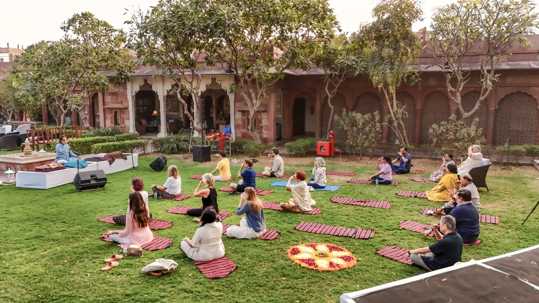 Ustad Nawab Khan leading morning naada yoga on the haveli lawn, guests seated on mats around a flower mandala, seen from above